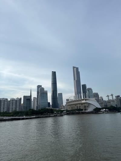This is a wide-angle, eye-level photograph of the Guangzhou skyline as viewed from across a body of water. The water in the foreground appears to be a river, with slight ripples on its surface and a dark, grayish-green color. A pedestrian walkway lined with trees is visible along the waterfront in the middle ground. Beyond the walkway, a dense collection of modern skyscrapers dominates the background.
Among the prominent buildings are the IFC (International Finance Centre) tower, a tall, slender skyscraper with a dark, reflective facade, and another very tall, angular skyscraper to its left. In the center of the skyline is a distinctive stadium with a white, curved roof structure supported by white beams. To the right of the stadium, there are several other modern buildings, including some with spire-like antenna structures. The sky above is overcast with scattered, wispy clouds, casting a soft, diffused light over the scene. The overall impression is a bustling urban landscape with a focus on modern architecture. This scene is consistent with the view of the Canton Tower and its surrounding skyscrapers in Yuexiu District, China.