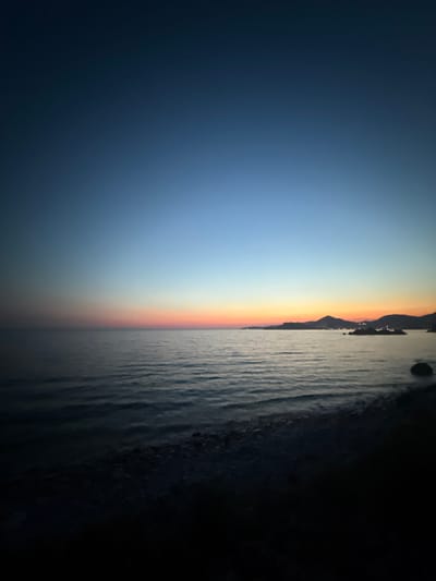 The image depicts a serene coastal scene at dusk, with the sky transitioning through shades of deep blue, orange, and pink near the horizon. The tranquil sea reflects the fading light, with gentle waves lapping at a pebble-strewn shore. In the distance, silhouetted hills rise against the colorful sky, dotted with faint lights suggesting a settlement. The scene is devoid of human presence. The location appears to be the coast near Sveti Stefan, Montenegro, known for its picturesque island and surrounding beaches.