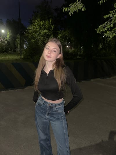 The image captures a young woman standing outdoors at night on a paved surface in Kazan, Russia. She has long, straight brown hair and is looking directly at the camera with a neutral expression. She wears a black long-sleeved cropped top with a quarter-zip neckline, blue denim jeans, small stud earrings, and a thin necklace. Her hands are positioned on or near her hips. In the background to her left, a low barrier displays alternating diagonal dark and light stripes, beyond which a bright streetlight illuminates trees and the dark sky. To her right and in the upper portion of the frame, dark foliage partially obscures the background. The ground is a dark grey asphalt or concrete surface, and the scene is predominantly lit by artificial light sources, creating areas of deep shadow.