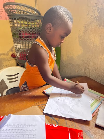 A young child with close-cropped dark hair is sitting at a wooden table in Rido, Nigeria, completing a worksheet. The child, who appears to be a toddler, is wearing an orange top with a stars and stripes design along the edges. He is holding a writing utensil in his right hand, poised to work on a paper covered in geometric shapes and numbers.
The table is cluttered with various papers, including a worksheet titled "THE ORIGIN OF COM". A red package, a box, and a stack of papers are also on the table. The child is sitting on a white plastic chair with cut-out designs. A gray laundry basket filled with clothing is visible behind the child. The wall behind the scene appears to be textured and painted in a muted color.