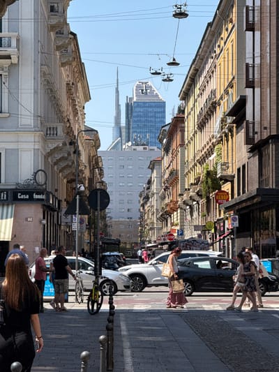 The image shows a street in Milan, Italy. The street is lined with buildings of varying heights, constructed in architectural styles including classical and modern. Pedestrians occupy the sidewalks on both sides of the street, including groups of people walking and individuals interacting with parked vehicles. Several cars are parked alongside the street. A bicycle is leaning against one of the vehicles. Street signage is visible on the sides of buildings. The sky is clear and the lighting appears to be mid-day sun. In the background, there are modern skyscrapers.