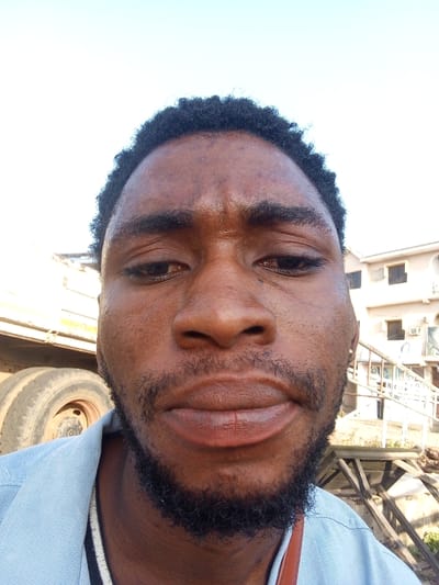 The image is a close-up, selfie-style photograph of a young Black man. He has dark, short, curly hair, dark eyebrows, and a beard. His skin tone is dark brown, and he has a light blue collared shirt on. The background suggests an outdoor, urban or semi-urban environment, possibly in the city of Lokoja, Nigeria. To the left of the man, a large, weathered tire and part of a vehicle, possibly a truck, are visible. Behind him and to the right, there is a building with multiple windows, and a wooden structure that appears to be scaffolding or a raised platform with a railing. The lighting indicates it is daytime, with a bright sky.