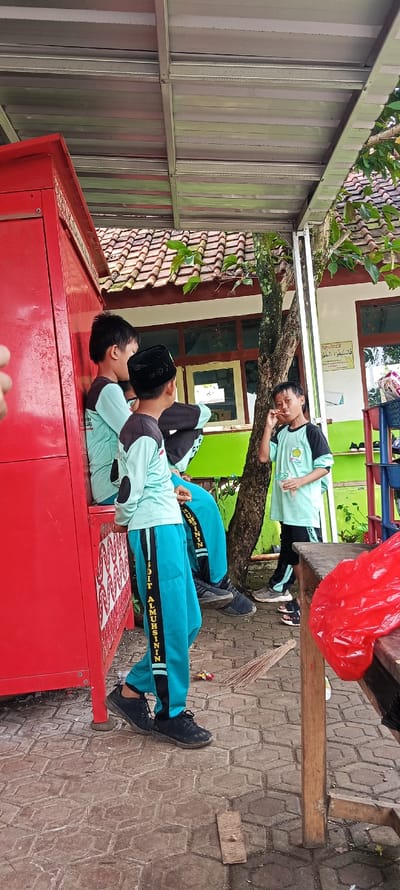 The image shows four young Indonesian boys standing near a red storage unit in Cangkuang, Indonesia, located at the coordinates -7.065 latitude and 107.551 longitude. The boys are wearing school uniforms and are interacting with each other, with one boy sitting on the storage unit while the other three are standing. The storage unit is made of metal and has a patterned design on the side. There is a large tree behind the boys, and there is a tiled walkway with brick-like patterns. A wooden table with a red plastic bag on it is in the background. The scene appears to be in a schoolyard. The boys appear to be school-aged and are likely students at the school.