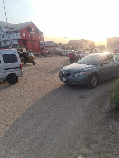 The image depicts a busy commercial street in Ajah, Nigeria, observed during a time of day with low sun angle, causing long shadows and strong lens flare in the upper right. In the left foreground, a silver panel van is parked or moving slowly, partially visible. Directly behind it, a yellow three-wheeled auto-rickshaw, or "keke," is present. On the right, a dark teal/green Toyota Camry sedan with a visible Nigerian license plate "LAGOS ABC-784FK" is positioned, facing slightly left. The immediate foreground shows a paved road surface with some wear and patches, and a narrow strip of grassy vegetation along the far right edge.
In the mid-ground, a red delivery motorcycle, distinguished by its large red insulated delivery box, is visible behind the Camry. Beyond the vehicles, a significant crowd of people is gathered, predominantly on the left side of the street near a prominent building. One individual within this crowd holds a white or light-colored umbrella. Numerous other vehicles, including cars and vans, are visible in the background, suggesting a congested area.
The background is dominated on the left by a multi-story building featuring a distinct red facade on its lower levels with multiple windows and some textual signage, topped by a lighter grey upper section. Various other commercial buildings and shops with readable but unidentifiable signs (e.g., "GLOBUS") line the street further down. Overhead utility poles and power lines are visible against the bright sky, indicating urban infrastructure. The overall scene reflects active vehicular and pedestrian traffic in a commercial district.