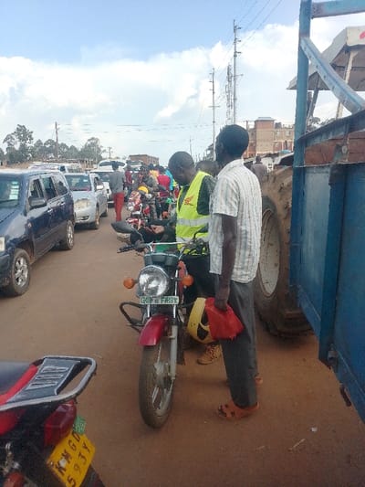 The image shows a busy street in Lwandeti, Kenya (latitude: 0.563, longitude: 34.805). There are several cars and motorcycles lined up in traffic, with a truck parked on the right side of the frame. Two men are standing near the front of the line of vehicles, one wearing a yellow reflective vest and the other in a striped shirt, holding a red bag. The man in the yellow vest is holding a phone and appears to be talking to the man in the striped shirt. The street is dusty and lined with buildings on one side. The sky is mostly cloudy.