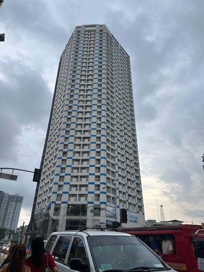 An overcast sky hangs above a tall, white, multi-story building with a checkered pattern of blue bands on its facade. The building, identified as the "Puso Towers" by text on its upper section, has numerous windows and balconies. Scaffolding is visible on the lower portion of the building, suggesting ongoing construction or maintenance. In the foreground, a white vehicle, possibly a taxi, is stopped in traffic. To the right of the taxi, a red jeepney, a common mode of public transportation in the Philippines, is also visible with passengers inside. Several people are seen in the foreground, some walking and others inside the vehicles. Street signs indicating "J.P. Laurel" are present. In the background, other buildings and a cell tower can be seen. The scene appears to be a street in Davao City, Philippines, given the street sign and the presence of a jeepney.