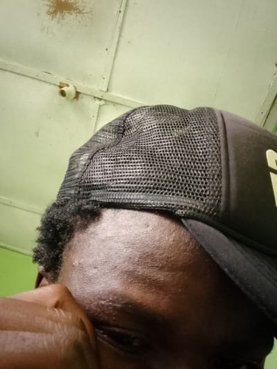 This media file is a close-up, low-angle shot of a person wearing a black trucker hat. The subject is positioned in the foreground, with only a portion of their face visible, including their forehead, part of their eye, and a hand resting near their face. The hat has a black mesh back and a solid front, with what appears to be a yellow logo on the side, though the text is not fully legible. The person has dark, curly hair peeking out from under the hat.
The background is a light green, textured surface, possibly a wall or ceiling, with visible seams or panels and a small, light-colored fixture attached to the upper part. There are some darker, irregular marks on the upper left portion of the background. The lighting suggests an indoor setting, possibly with artificial light.
The image appears to be a selfie, taken from a very low angle, looking upwards towards the subject and the ceiling. The visible portion of the face does not convey a strong emotion, appearing neutral. The overall impression is candid and informal. The location context provided is Jauro Yinu, Nigeria.