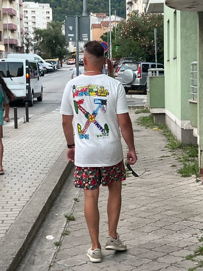 In Budva, Montenegro, a man with a fade haircut walks away from the camera on a paved sidewalk. He is wearing a white t-shirt with colorful graphics and floral patterned shorts. He holds a pair of sunglasses in his right hand. To his left, a drain runs along the edge of the sidewalk, separating it from a grassy area. Across the street, vehicles are parked and moving, including a white van and a silver SUV. Buildings line the street, with apartment buildings visible in the background. The overall scene depicts a street in a European coastal town.