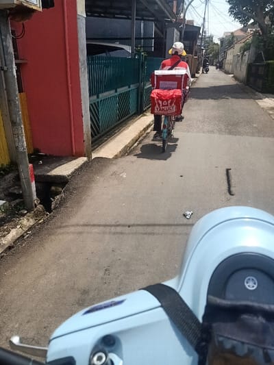 A Yakult vendor rides a bicycle on a narrow street. The vendor wears a white hat and red shirt, with delivery containers labeled "Yakult" attached to the bike's rear. The street is lined with houses, and there is a scooter in the foreground with a black strap across the handlebars. The road surface is asphalt. The photo is taken at eye level from the scooter.