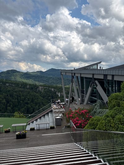 This media file shows an outdoor seating area on a multi-level structure overlooking a densely forested mountain range. The scene is set during the day, with a sky filled with dramatic, grey clouds, hinting at potentially overcast or changeable weather.

The foreground is dominated by a wooden deck with several tables and chairs, suggesting a restaurant or café. Some tables are occupied by people, who appear to be dining or socializing. There are also potted plants, some with colorful flowers, adding decorative elements to the space. A glass railing separates the deck from the steep drop-off, offering an unobstructed view of the mountains.

The architecture of the building is visible, featuring a modern design with large glass windows and prominent steel supports. Behind the seating area, lush green trees cover the mountainside, creating a vast, natural landscape. The time of day appears to be late afternoon or early evening, judging by the lighting.

The location is identified as Adlerskiy, Russia. The overall mood is one of relaxed enjoyment, with people taking in the scenic views from the elevated vantage point. There is no visible text within the image.