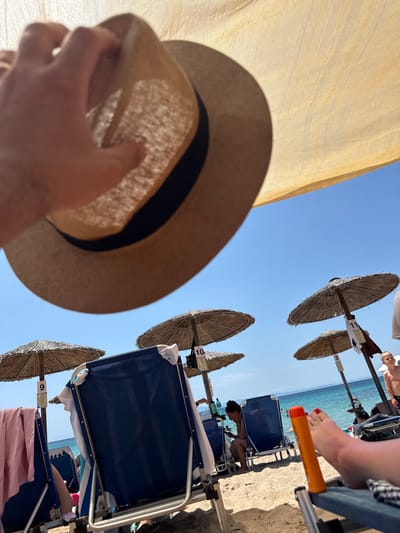 A first-person perspective shot of a person holding a straw hat on a sunny beach in Potos, Greece. The image is oriented vertically. The bright sun is casting shadows from beach umbrellas onto the sand. Several thatched umbrellas and blue beach chairs are visible. In the background, the turquoise water of the sea meets a clear blue sky. Some people are relaxing on the beach chairs, and a hand with red nail polish is visible holding an orange sunscreen tube. Numbers are visible on the beach umbrellas, indicating chair or umbrella numbers. One umbrella has the number "11", another "3", and one "10". Another umbrella shows the number "9". The overall mood is relaxed and summery.