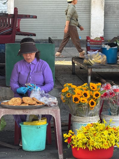 In Nha Trang, Vietnam, a woman in a purple shirt and a dark visor is seated behind a table, preparing food. She wears blue gloves and appears to be working with small items in her hands, possibly seasonings or ingredients. In front of her, on a silver tray, are several circular, golden-brown food items. Beside the tray are clear plastic bags containing more of these items. A large turquoise bucket is visible beneath the table, partially filled with what appears to be a yellow plastic bag.
In the background, a person wearing a light green hooded sweatshirt and brown checkered pants walks away from the camera. To the right of the scene, a variety of flowers are displayed. A bouquet of sunflowers is in a clear plastic wrapper, and next to it are red flowers, also wrapped. In front of these, a large red basin overflows with small yellow flowers. A metal table with red crates stacked on it is situated behind the seated woman, with a bag of bananas visible on the table. The environment suggests a street market setting, with a closed metal roll-up door in the background.
