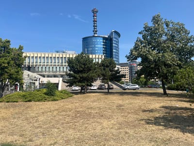 A daytime, sunny shot captures the exterior of a modern building complex in Budapest, Hungary. The foreground is dominated by a dry, mowed lawn, with a few scattered pieces of litter. Behind the lawn, a multi-story building with a light-colored facade and numerous windows stretches across the midground. Tall green trees flank the building, and a wide set of concrete stairs leads up to the entrance.
To the right of this building, a taller, curved building with a sleek blue glass exterior rises. Atop this structure is a distinctive, tall communication tower with a segmented design, featuring red and white sections and several platforms. Further in the background, other modern buildings can be seen, suggesting an urban environment. Several cars are parked in front of the buildings, and a few people are visible near the entrance of the lighter-colored building, one of whom is standing by a decorative sculpture. The sky is a clear, bright blue. The overall scene conveys a sense of a clear, warm day in a city. Visible text on the building reads: "VÁR ALLATVÉDELEM".