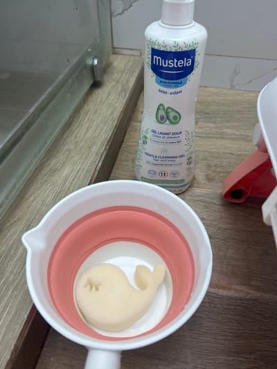 A close-up, eye-level shot captures a bathroom scene on a wooden countertop. In the foreground, a white and pink collapsible basin holds a light yellow, whale-shaped sponge. To the right of the basin, a tall white bottle of Mustela Gentle Cleansing Gel is positioned upright. Behind the basin and bottle, a portion of a glass shower enclosure with a silver handle is visible. The background features a tiled wall. The overall impression is one of a well-organized, clean, and functional bathroom space. No specific landmarks or indications of Plovdiv, Bulgaria are present in the image.