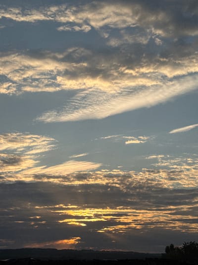 This is a landscape photograph taken at sunset in Nizhnyaya Gar', Russia. The sky is filled with layers of clouds, some dark and brooding, others tinged with gold and orange from the setting sun. Rays of light pierce through the clouds, creating dramatic sunbeams. The horizon is lined with a dark silhouette of trees and what appears to be a dark structure in the foreground. The overall atmosphere is one of serene, natural beauty.