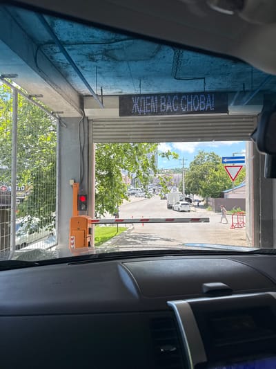 The image shows a view from inside a vehicle, looking out through an open gate. Above the gate, a digital sign displays the Cyrillic text "ЖДЕМ ВАС СНОВА!" which translates to "WE ARE WAITING FOR YOU AGAIN!". To the left, a traffic light shows a red signal. An orange automatic barrier arm is in the lowered position. Outside the gate, there is a street with several vehicles, including cars and vans. A person is walking across the street, and another person is standing near some red temporary barriers. A yield sign and a blue directional sign with a white arrow pointing left are visible on the right side of the street. Lush green trees line the street, and buildings can be seen in the background, suggesting an urban or suburban setting. The overall scene appears to be an entrance or exit to a parking area or a facility with controlled access. The image was taken in Novorossiysk, Russia.