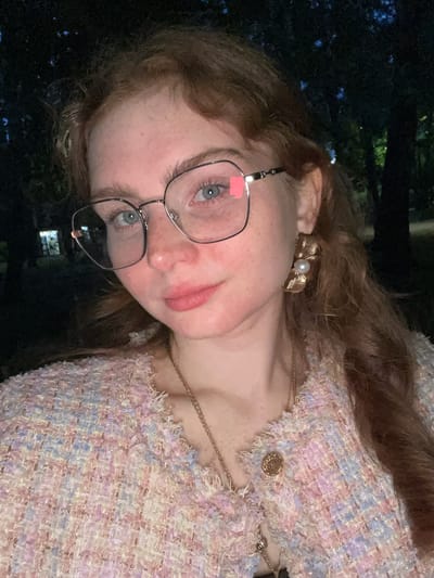 A young adult female with reddish-brown, wavy hair and fair skin, showing visible freckles on her nose and cheeks, is featured in a close-up photograph. She has light blue eyes and is wearing large, thin-framed metal glasses; a small pink rectangular adhesive note or sticker is affixed to the upper right portion of her right lens. Her lips are subtly tinted pink. She is dressed in a textured tweed jacket or top, characterized by a woven fabric incorporating pastel shades of pink, blue, yellow, and white, with intentionally frayed edges along the neckline. A thin gold-colored chain necklace with a small, circular, ornate gold pendant is visible on her chest. A prominent gold-colored earring, designed in a fanned or shell-like shape with a central white pearl, is worn in her right ear. The background is dark and out of focus, revealing the silhouettes of trees and faint, blurred artificial lights, indicating an outdoor, nighttime or late dusk setting. The subject is well-lit, suggesting direct artificial illumination, likely from a camera flash. The photograph is stated to be taken in Kazan, Russia.