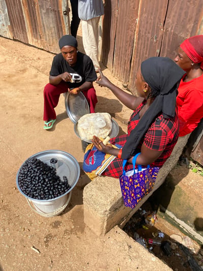 In Bukuru, Nigeria, three individuals are engaged in a transaction at an outdoor market. A woman in a black beanie and red pants is seated, holding a piece of food and a metal tray. Next to her, another woman in a black headscarf and a red and black plaid dress is pointing, seemingly in conversation with the first woman. A third woman, wearing a red headscarf and a red shirt, stands behind them. In the foreground, a large metal basin filled with dark, oval-shaped fruits sits on a white bucket. A smaller metal dish on top of the basin contains a few of the same fruits and a small metal container. Behind the seated women, a metal structure with vertical panels forms the backdrop. A concrete structure runs along the right side of the image, with a ditch filled with debris next to it.