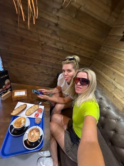 Two women are sitting on a brown leather couch in a rustic setting, likely a cafe with wooden paneled walls. One woman, in the foreground, wears a bright green t-shirt and dark shorts, her legs crossed. She is wearing large, reflective sunglasses and her blonde hair is down. The other woman, behind her, wears a white t-shirt and has her blonde hair pulled up in a bun, also wearing sunglasses pushed up on her head. She sticks her tongue out playfully at the camera.
On a wooden table in front of them is a blue tray with a breakfast or snack. The tray holds two cups of coffee with latte art, two pieces of toasted bread, a small dish of what appears to be tuna or pâté, and two small dishes of red condiments, possibly jam or tomato sauce. A dark menu is also visible on the table.
One of the women is holding a smartphone, taking a selfie or a photo of the scene. The lighting suggests it might be daytime, possibly afternoon. The overall mood appears to be relaxed and fun. The location context provided is València, Spain. No legible text is visible in the image.
