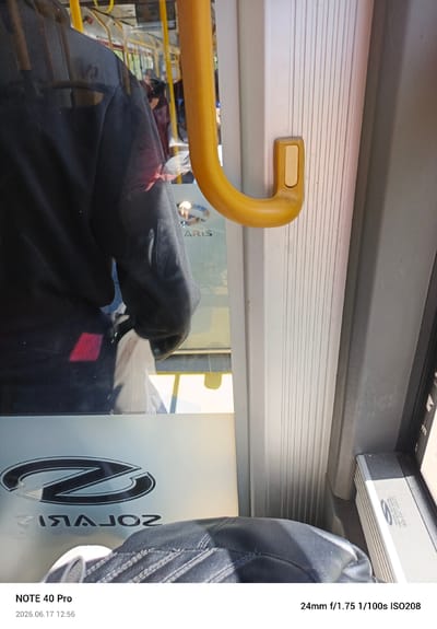 The image shows the interior of a public transport vehicle, possibly a bus or tram, in Warsaw, Poland. In the foreground, a portion of a black leather seat is visible. The focus is on the right side of the frame, where a yellow handrail is prominent. To the right of the handrail, a white textured panel with vertical ridges runs along the wall. A small, rectangular beige button is mounted on this panel. Through a window to the left, reflections reveal passengers and the interior of the vehicle, including yellow support poles and seats. A prominent "Z" logo is reflected on the glass surface, appearing reversed as "ZIAJOZ". The bottom of the image displays text indicating "NOTE 40 Pro" and the date "2026.06.17 12:56", along with camera settings "24mm f/1.75 1/100s ISO208".