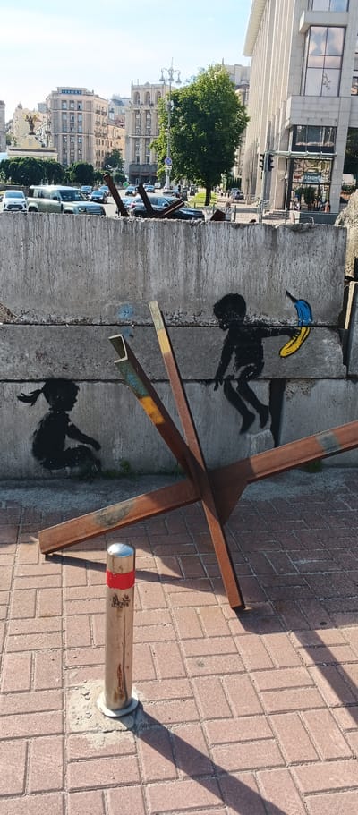 In Kyiv, Ukraine, a concrete barrier, likely part of a defensive structure, is spray-painted with two black silhouettes of children. On the left, a child with a ponytail is crouched down. On the right, a child is depicted jumping and holding a yellow and blue object, symbolizing the Ukrainian flag. In front of the barrier, a "Czech hedgehog" anti-tank obstacle made of rusted metal beams is positioned. A silver bollard with red and white markings stands in the foreground on a red brick paved sidewalk, casting a long shadow. In the background, city buildings and a tree are visible, along with traffic and pedestrians.
