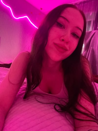 In a dimly lit room, a young woman with long, dark hair and prominent eye makeup lies on a pink bedspread. A neon pink light fixture snakes along the wall behind her, casting a vibrant glow over the scene. Her face is tilted towards the camera, with a slight smile playing on her lips. The room appears to be a bedroom, with a curtained window visible in the background. The overall impression is one of intimacy and a youthful, playful atmosphere. The image has not been geolocated to Votkinsk, Russia.