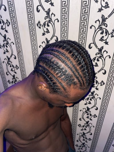 An angled close-up captures a man with dark skin and braided hair, seen from the neck up. The braids are intricate and run across the top of his head. He is shirtless, revealing his chest and upper body. The background features a wall with a repeating black and white geometric and floral pattern. The lighting appears artificial, casting a subtle sheen on his skin and hair.