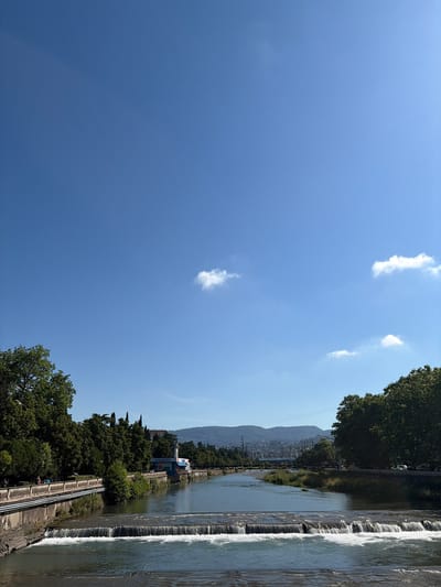 This image captures a scenic view of the Matsesta River in Sochi, Russia, under a clear blue sky dotted with a few wispy clouds. The river flows from the distance, creating a small waterfall or cascade in the foreground where the water churns over a series of rocky ledges. The banks of the river are lined with lush green trees, indicating a temperate climate.
On the left bank, a decorative stone embankment with an ornate railing runs alongside the river. A pathway is visible on top of this embankment, and several people are casually strolling along it, enjoying the outdoors. Further along the river, on the right bank, a modern-looking building with blue and white accents stands out, possibly a cafe or a public facility, with some signage visible on its facade. The background reveals rolling hills or mountains, suggesting the city's proximity to natural landscapes. A bridge spans the river in the middle distance, and further back, more bridges and structures hint at the urban environment of Sochi. The overall atmosphere is peaceful and idyllic, showcasing a harmonious blend of natural beauty and urban development.