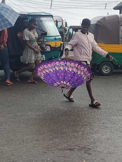 A person in a pink jacket and black beanie holds a vibrant purple umbrella with a floral pattern. They are standing with one leg extended, mid-stride, on a wet, grey asphalt road. The person wears black slides with white soles. Behind them, a green and yellow auto-rickshaw, a common mode of transport, is visible. Further back, another auto-rickshaw is parked. Two women are seated inside one of the auto-rickshaws. One woman, wearing a black and white patterned dress, holds a smartphone. A person in a red top and blue jeans lies down, partially covered by a blue and white checkered umbrella. The scene suggests it is raining, as the ground is wet and umbrellas are in use. The sky appears overcast, indicating a cloudy day, likely daytime due to the presence of auto-rickshaws and people active in the street. The overall atmosphere is a typical street scene in Gwong, Nigeria, during wet weather.