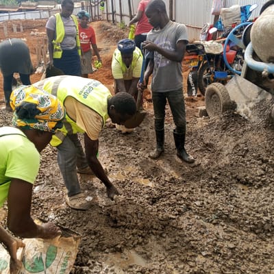 The image depicts a muddy construction site, possibly in Borabu Chitago ward, Kenya, based on visual context and implied details from limited visible text. Several individuals are engaged in labor, including mixing cement. Some wear yellow safety vests, while others are in casual attire. One person wields a shovel, and another appears to be opening a bag labeled "CEMENT." A cement mixer and a motorcycle are partially visible, suggesting their use in the construction process. The background shows unfinished structures and scaffolding. The overall environment is unpaved, with muddy terrain dominating the foreground.