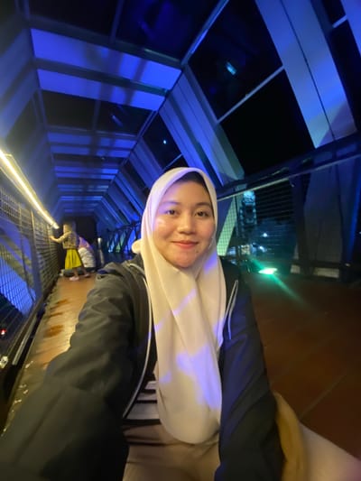 The image shows a woman wearing a light-colored hijab and a black jacket, taking a selfie. She is in a long, narrow corridor with blue lighting on the left and a wooden floor on the right. The corridor appears to be part of a building or a walkway with metal railings on both sides. In the background, another person is visible further down the corridor, wearing a leopard-print jacket and a yellow skirt. The lighting suggests it is nighttime or an indoor environment. The woman's expression is a soft smile. The location is identified as Banjarbaru, Indonesia.