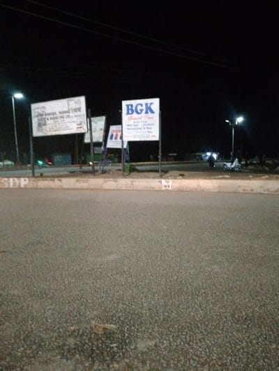 This is a nighttime photograph taken in Bukuru, Nigeria, showing a street scene with several signs and a sidewalk. The lighting suggests it's after dark, with artificial lights from street lamps and businesses illuminating the area. The ground is paved, and there's a concrete barrier or curb in the foreground.
Two prominent signs are visible. One, on the left, is a white sign with dark text, appearing to advertise services and training, possibly related to marketing. The text is partially obscured and somewhat blurry, but "SERVICES, TRAINING CENTRE" and "& MARKETING LTD." are discernible. To the right of this sign is another white sign with bold blue lettering that reads "BGK". Below this, smaller text lists various services, including "Guest House", "Mini Hall", "Chairs", and "Restaurant & Bar".
Further down the road, other lights and structures are visible, including what appears to be a street light and some buildings in the distance. A white motorcycle is parked on the side of the road, and a person is walking in the background near some lights. The overall mood of the image is one of a typical evening in a town, with businesses open and people moving about. The weather appears clear, as the night sky is dark with no visible clouds.