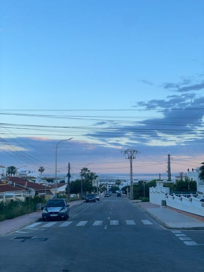 The image depicts a street scene in Rojales, Spain. The street is paved with asphalt and has white lines indicating lanes and pedestrian crossings. A dark-colored car is parked on the side of the road, facing the direction of traffic. Several other cars are visible further down the street. White buildings with flat roofs line the side of the road, interspersed with palm trees and other vegetation. Utility poles and power lines are visible against the sky, which is a gradient of blue and pink hues, indicating either sunrise or sunset. The overall scene suggests a quiet residential area near the coast, as the sea is faintly visible in the background.