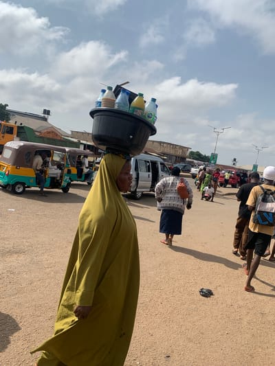 A woman walks in Gura Top, Nigeria, with a large black basin filled with plastic bottles of drinks balanced on her head. She is wearing a flowing olive green garment that covers most of her body and a headscarf. The scene is an outdoor market or street with several vehicles visible, including three-wheeled auto-rickshaws (keke napep), vans, and cars. Other people are walking in the background, some carrying bags. The ground is paved with a light-colored, sandy material. The sky is partly cloudy.