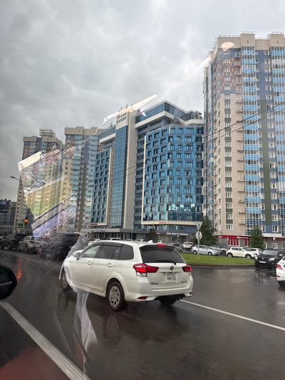 The image depicts a street scene in Krasnoyarsk, Russia, characterized by modern, multi-story buildings and traffic. In the foreground, a white station wagon is driving on a wet road, reflecting the surrounding environment. The buildings are tall, with facades featuring a combination of glass panels, colored bricks, and concrete. The sky is overcast, suggesting cloudy weather. Several other vehicles are visible on the road and parked along the side. The overall impression is that of an urban environment with ongoing traffic flow.
