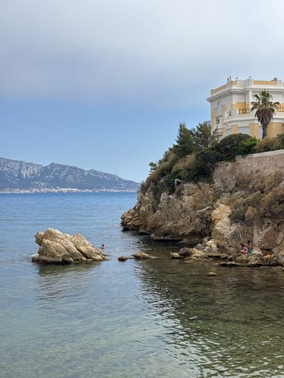 The image shows a view of the Mediterranean Sea in Marseille, France, with a rocky coastline in the foreground and a city skyline in the distance. On the right side of the image, a pale yellow and white building sits atop a cliff, surrounded by lush green trees and shrubs. The building has a balcony with yellow railings and a palm tree is visible to its right. The rocky cliff face is textured and shows signs of erosion and vegetation growth. In the water, there are several large rocks. On one of these rocks, a person is swimming. Further to the right, on the rocky shore, two people are sitting, one wearing a pink bikini and the other wearing a green garment. The water is clear and calm, with sunlight reflecting off its surface. In the background, a chain of mountains stretches across the horizon, with a city built along its base. The sky is a light blue with a few wispy clouds. The overall scene depicts a serene coastal landscape with elements of both nature and human habitation.