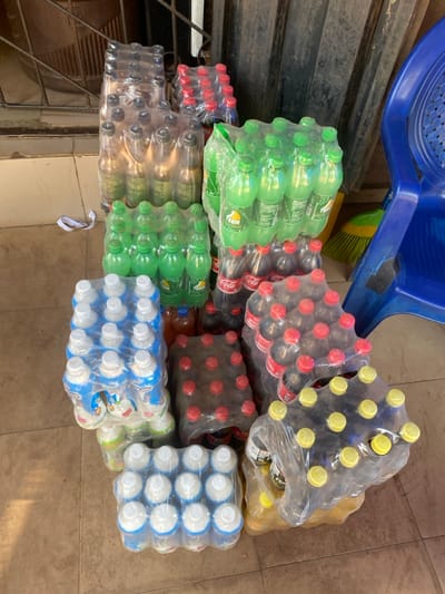 The image displays a ground-level view of numerous commercially packaged beverages stacked on a light brown tiled floor within a commercial or storage area in Okura, Nigeria. The scene is dominated by multiple shrink-wrapped plastic bottle packs, including green-labeled sodas (some branded "Teem"), dark-liquid sodas with red caps (branded "Coca-Cola"), various other dark-capped beverages, yellow-liquid beverages with yellow caps, and white/blue-labeled dairy or yogurt drinks with white caps (one partially visible label appears to be "Hollandia"). Most packs contain multiple bottles, typically arranged in arrays of 6, 9, or 12.
To the right, a blue plastic monobloc chair is partially visible, behind which a yellow broom with a brown handle is propped against a dark surface. The background features a dark, possibly corrugated metal wall or door, with a metal grille visible in the upper left. A small, crumpled white piece of material lies on the floor near the top-left stack of beverages. No individuals are present, and no actions or interactions are depicted.