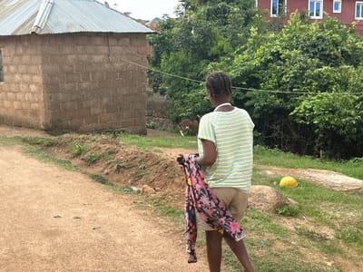 In Gwong, Nigeria, a young person stands with their back to the camera, holding a colorful, floral-patterned garment. They are wearing a light gray and neon green striped t-shirt and light brown shorts. The scene is outdoors, with a dirt path in the foreground. To the left, a simple building constructed of cinder blocks with a corrugated metal roof is visible. Further back, lush green trees and bushes dominate the landscape, with a glimpse of a red building with white-framed windows in the distance. A few goats are present in the background, one near the building and another lying down in the grassy area. A yellow object, possibly a large bowl or basin, sits on the grass in the middle ground. The lighting suggests it is daytime, likely overcast or late afternoon, given the soft shadows and diffused light. The overall impression is one of a rural or semi-rural setting in Nigeria.