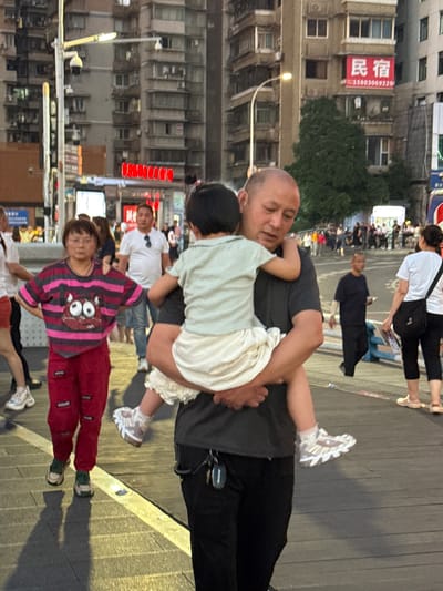 The image depicts an urban street scene at dusk or early night in Yuzhong District, China. The central focus is a man with a shaved head, dressed in a dark shirt and pants, carrying a young child in a light-colored top and white ruffly shorts. Another child's head is partially visible near the man's left hip. In the foreground, a woman with short dark hair is walking, wearing a distinctive pink and grey striped long-sleeved shirt with a large cartoon-like face graphic, paired with red pants and green-banded sandals.
The background is dominated by multi-story buildings, characteristic of a city environment. Many windows are visible, and streetlights are illuminated, casting shadows on the dark-toned pavement. Several commercial signs are mounted on the buildings. A prominent red rectangular sign on one building clearly displays the Chinese characters "民宿" (Mínsù), meaning "Guesthouse" or "Homestay," followed by a phone number "15803069129." Other blurry signs with Chinese text are also visible further down the street. People are scattered throughout the scene, suggesting a casual evening outing or pedestrian activity. The overall mood appears relaxed and ordinary for a city street.