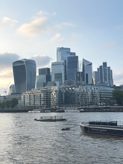 This image depicts a view of the city of London, United Kingdom. Several modern skyscrapers dominate the skyline, including the "Walkie Talkie" building. The River Thames is visible in the foreground, with a large boat, marked "Port of London Authority," on the water. The scene appears to be captured during the daytime, with a partly cloudy sky.