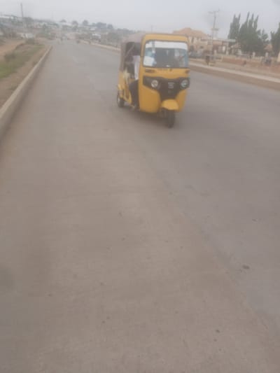 A yellow auto-rickshaw is driving on a paved road in Sabon Gari, Nassarawa, Nigeria. The auto-rickshaw has a covered passenger area and a driver. The driver is wearing a white shirt. There is at least one passenger in the auto-rickshaw. The road is a two-lane highway with a concrete barrier on the left side. In the background, there are buildings, trees, and a utility pole. The sky is overcast. The image is taken from a slightly elevated perspective.