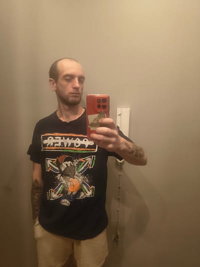 This media file captures a selfie taken by a man in what appears to be a bathroom or a room with plain walls, likely indoors. The man is the primary subject, wearing a dark t-shirt with a graphic design on it. The design features a cartoon character holding a basketball, surrounded by stylized arrows and splashes of paint. The text "POWDER" is also visible on the shirt in a bold, stylized font. He has short dark hair, stubble, and several tattoos visible on his arms. He is holding a red smartphone, angled to take the picture, and a US dollar bill is partially visible within the phone case.
The image is oriented sideways, suggesting the photo was taken with the phone held in a portrait orientation, but the image file itself has been rotated. The lighting is dim, casting shadows on the wall behind him. There is a white electrical outlet cover and a coiled white wire visible on the wall in the background. The overall impression is that of a casual, personal photograph. No specific time of day or weather conditions are indicated. The location context provided is Grójec, Poland, but there are no visual cues within the image to confirm this specific location. The man's expression is neutral.