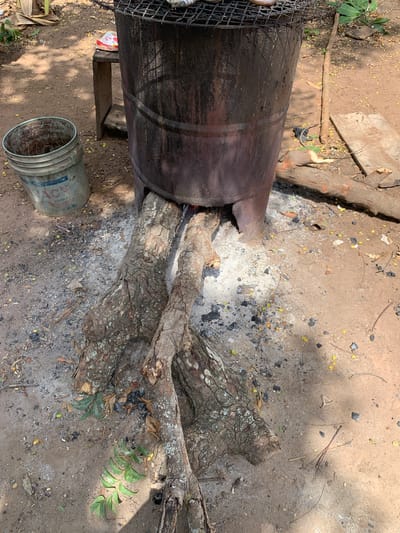 The image depicts an outdoor scene in Ilorin, Nigeria. A large, rusty, metal drum with a portion cut out is positioned horizontally. Logs are placed inside the drum, apparently for burning, with evidence of ash and charred remnants scattered around the base. To the left of the drum, a wire mesh structure is partially visible. In the lower-left corner, a used bucket is lying on its side. The ground is dirt, with scattered leaves, debris, and small yellow fruits. Patches of sunlight filter through the overhead canopy, creating shadows across the scene. No people are visible in the image.