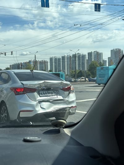 A silver Hyundai Solaris sedan with a severely damaged rear bumper and trunk is stopped at an intersection in Saint Petersburg, Russia. The license plate reads "K449PB147". A blue bus is visible in the background, along with multi-story residential buildings. The sky is cloudy. The image appears to have been taken from inside another vehicle.