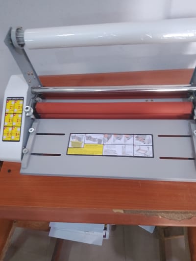 This is a close-up, eye-level, interior shot of a laminating machine placed on a wooden desk. The machine is white and gray, with a roll of clear laminating film loaded at the top and two red rollers visible below. To the left of the machine is a vertical panel with yellow instruction labels. To the right of the rollers, there is a gray surface with two horizontal slots and another instruction label featuring several small diagrams. The desk is made of polished wood and appears to be a standard office or home desk. The background is a plain, light-colored wall. There are no people or any other objects in the frame besides the laminating machine and the desk it rests upon. The image does not provide enough information to determine its specific location within Jalingo, Nigeria.