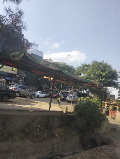 This daytime photograph captures an outdoor scene in Nairobi, Kenya, showcasing a combination of urban infrastructure and natural elements under a bright, sunny sky.

In the immediate foreground, a rough concrete retaining wall or drainage ditch with earth and sparse vegetation is visible, including a concrete culvert. Above this, a bustling unpaved parking area is filled with numerous parked vehicles, predominantly cars and SUVs of various models and colors.

A prominent feature in the mid-ground is a long, rectangular canopy structure, supported by metal poles and covered with weathered fabric in shades of green, orange, and brown. Underneath this canopy, towards the right side of the frame, a person wearing a yellow shirt and dark trousers can be seen, appearing to face away from the camera.

The background is populated with several multi-story buildings and lush green trees. On a building to the left, a blue sign with white text partially reads "SAMBU PATIENT CENTRE." Further to the right, on another building, a white sign with black lettering clearly displays "L.V. MACHAKO & CO. ADVOCATES."

The sky is light blue with scattered white clouds, indicating clear weather. A small bird is distinctly visible in flight in the upper right portion of the sky. The overall impression is that of an active commercial or residential area within the city.