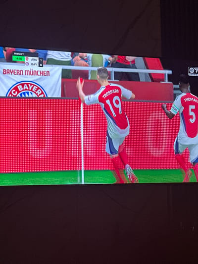 The image shows a screenshot from a soccer video game, displayed on a television screen. In the game, two players wearing red and white kits are visible on a field. One player, identified as "TROSSARD 19" on his jersey, is celebrating a goal, raising his arm. Another player, "THOMAS 5", is to his right, also in a celebratory pose. A score overlay in the top left corner shows "FRIENDLY 14:10 ARS 1 BAY 0". A banner with "BAYERN MÜNCHEN" and a club crest is visible in the background. The scene is illuminated by the bright display of the television. The image is taken from a slightly elevated angle, looking down at the screen. There is no discernible connection to Gwarinpa, Nigeria.
