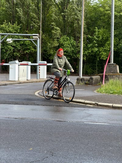 A middle-aged person on a black bicycle is stopped at a pedestrian crossing gate in Esch-sur-Alzette, Luxembourg. The person is wearing a red head covering, glasses, a green jacket, grey trousers, and brown and orange striped leg warmers. They also have brown shoes and green gloves. The crossing gate is composed of white and red barriers and is adjacent to a concrete barrier. Lush green trees are in the background. The road is wet and asphalt.