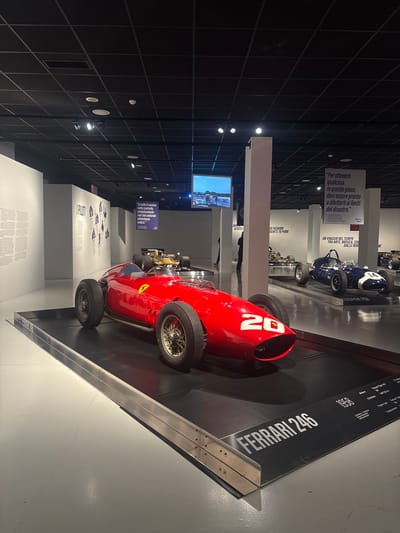 This media file captures an indoor exhibition space, likely a museum in Turin, Italy, showcasing vintage race cars. The image is rotated 90 degrees to the left, showing a red Ferrari 246 race car prominently displayed on an elevated black platform in the foreground. The car has "20" painted in white on its nose and dates to 1958, as indicated by a large display panel next to it. This panel also lists details such as "Engine type Ferrari Tipo 143 V6" and "Displacement 2417.33 cc".
Behind the red Ferrari, a blue Cooper T51 race car from 1959 is visible on a similar platform. The background features dark walls with grid patterns and track lighting. White display pillars and panels are arranged throughout the space. One large banner displays an Italian quote: "Per ottenere qualcosa in questo gioco, devi essere pronto a dilettarti ai limiti del disastro." (To achieve anything in this game, you must be prepared to dabble in the boundary of disaster), attributed to Stirling Moss. Another banner on the far left reads: "Se tutto ti sembra sotto controllo, semplicemente non stai andando abbastanza veloce." (If everything seems under control, you're just not going fast enough.) Further textual displays include "PILOTI" (Pilots) and "UN VIAGGIO NEL TEMPO TRA ARTE, MUSICA, CINEMA" (A journey through time between art, music, cinema). A digital screen embedded in a pillar shows a blurred image of a landscape or cityscape. The setting suggests a well-curated historical exhibit, celebrating automotive racing and its philosophy. Faint figures of visitors are visible in the background, viewing the exhibits.
