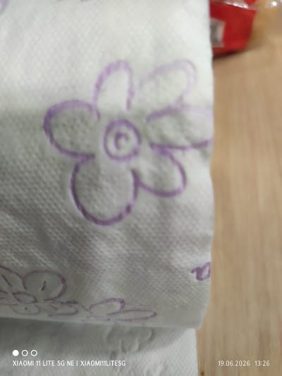 The image is a close-up, eye-level shot of a roll of white toilet paper with a subtle embossed floral pattern in purple. The pattern appears to be a stylized flower with multiple petals and a circular center. The toilet paper is on a wooden surface, possibly a table or counter, which is out of focus in the background and exhibits a warm, light brown tone. The lighting appears to be artificial, casting a soft, even glow. In the bottom right corner of the image, a timestamp is visible: "19.06.2026 13:26". In the bottom left corner, camera information is displayed: "XIAOMI 11 LITE 5G NE | XIAOMI11LITE5G". The image does not contain any identifiable landmarks or clues to suggest a specific location within the city of Tyumen, Russia. The scene is static and focuses solely on the product and its immediate surroundings.